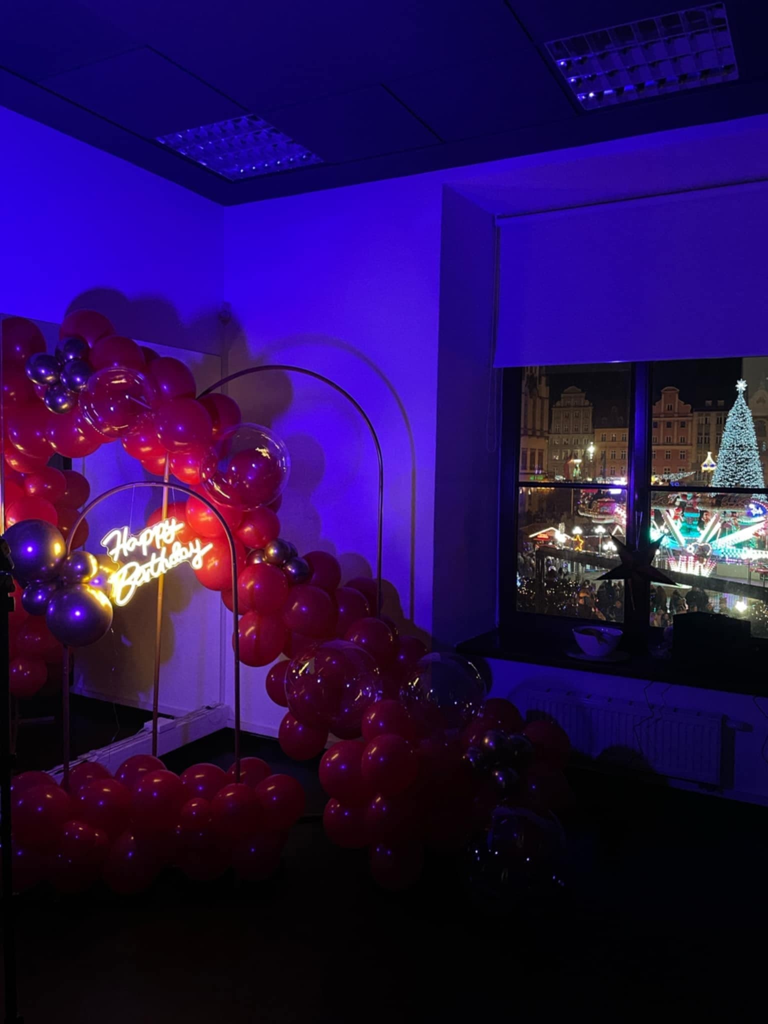 Ta sama girlanda balonowa w kadrze z oknem, za którym widać oświetlony choinką plac miejski nocą